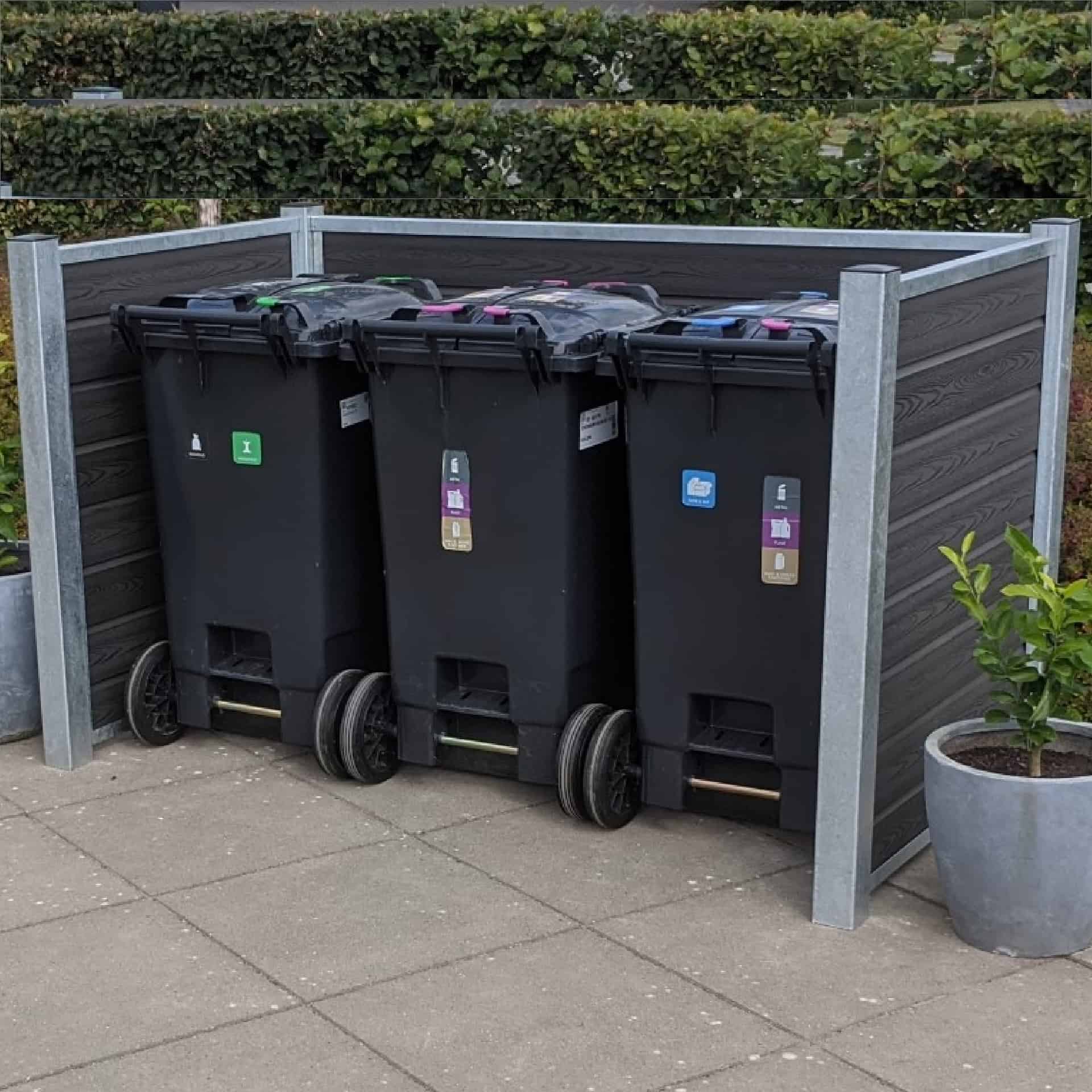 Et sæt med tre sorte skraldespandsspande, hver med et forskelligt farvet låg og etiket, er placeret i en pæn træ-skraldespandsskjul udendørs. Beholderne hviler på en flisebelagt overflade med potteplanter i nærheden og grønne hække i baggrunden, alt sammen understøttet af robuste stålstolper.