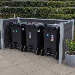 Tre sorte skraldespande på hjul med farvede etiketter, der angiver genbrugstyper, er opstillet på en overskuelig måde i en Sort komposit skraldespandsskjul, sikret af stålstolper. Skraldespandene står på en brolagt overflade ved siden af pænt klippede hække og en potteplante.
