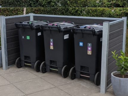 Tre sorte skraldespande på hjul med farvede etiketter, der angiver genbrugstyper, er opstillet på en overskuelig måde i en Sort komposit skraldespandsskjul, sikret af stålstolper. Skraldespandene står på en brolagt overflade ved siden af pænt klippede hække og en potteplante.