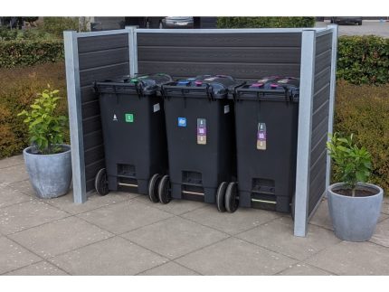 Tre sorte skraldespande på hjul er placeret side om side på et flisebelagt udendørsareal, omsluttet af et plankeværk med stålstolper på tre sider. Skraldespandene har farvede låg til genbrug. To store, runde plantepotter med grønne buske er placeret på hver side af slagsen Komposit skraldespandsskjul.