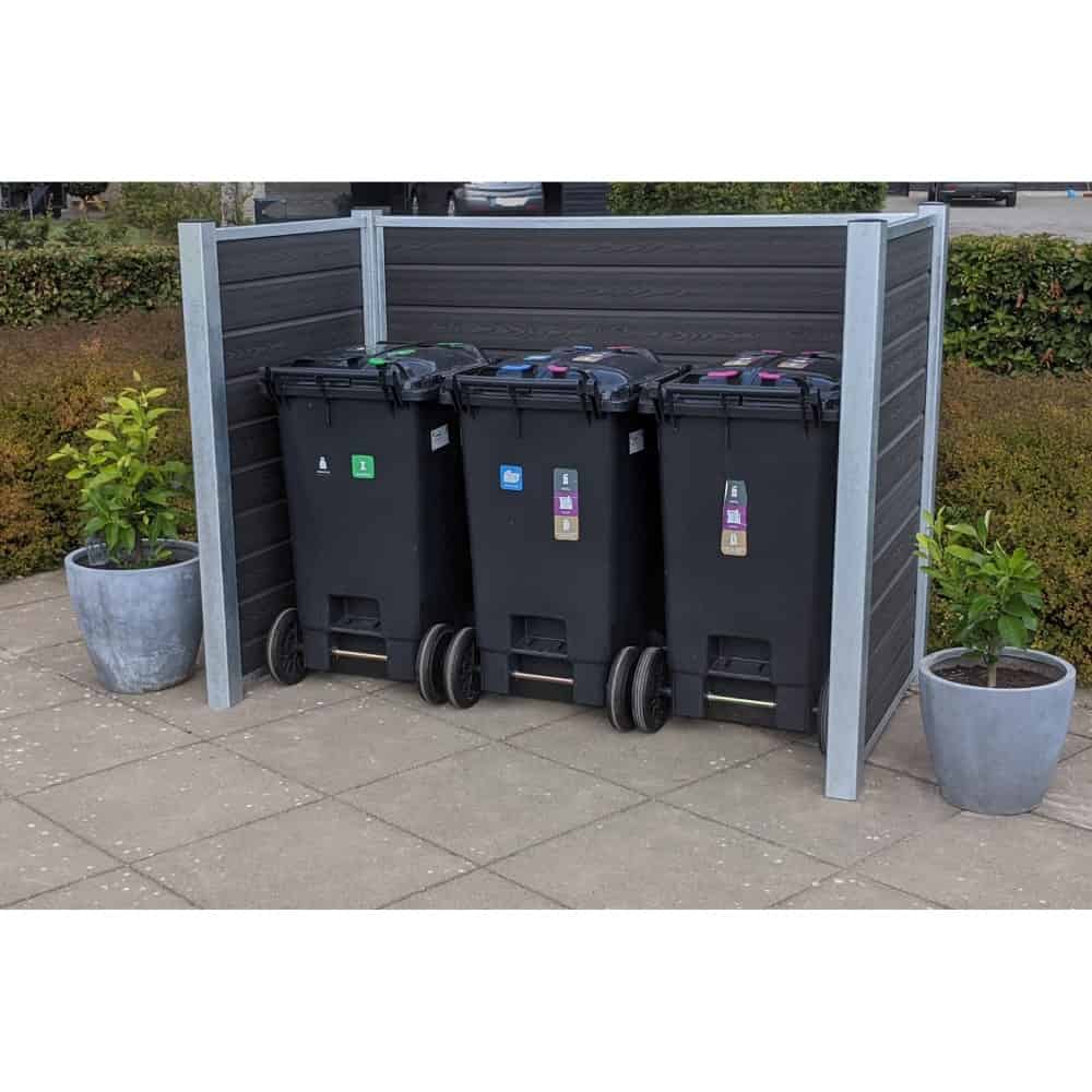 Tre sorte skraldespande på hjul er placeret side om side på et flisebelagt udendørsareal, omsluttet af et plankeværk med stålstolper på tre sider. Skraldespandene har farvede låg til genbrug. To store, runde plantepotter med grønne buske er placeret på hver side af slagsen Komposit skraldespandsskjul.