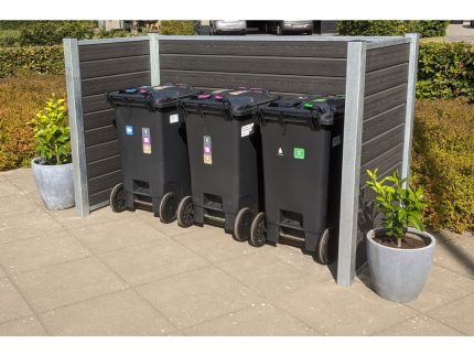En række af tre sorte skraldespande på hjul er placeret på en asfalteret overflade. Hver beholder har forskellige farvede låg og etiketter til at sortere affald. Komposit skraldespandsskjul indhegningen, forstærket med stålstolper, inkluderer to potteplanter på hver side. Buske er i baggrunden.