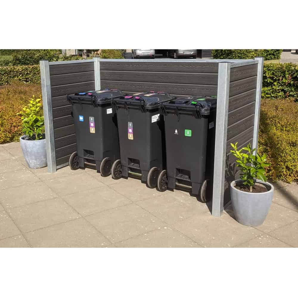 En række af tre sorte skraldespande på hjul er placeret på en asfalteret overflade. Hver beholder har forskellige farvede låg og etiketter til at sortere affald. Komposit skraldespandsskjul indhegningen, forstærket med stålstolper, inkluderer to potteplanter på hver side. Buske er i baggrunden.