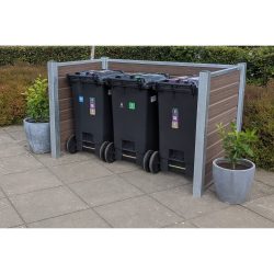 Et affaldshåndteringsområde med tre sorte skraldespande på hjul er omsluttet af et beskedent plankeværk understøttet af stålstolper. To store grå plantepotter med grønt løv sidder på hver side af skraldespandene. Rammerne er en udendørs terrasse med en hæk i baggrunden.