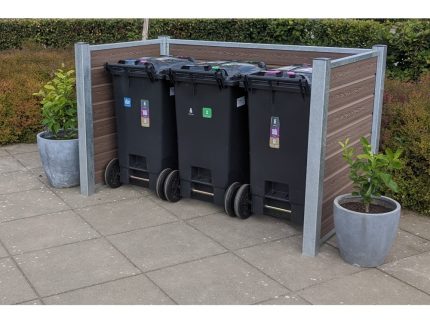 Et affaldshåndteringsområde med tre sorte skraldespande på hjul er omsluttet af et beskedent plankeværk understøttet af stålstolper. To store grå plantepotter med grønt løv sidder på hver side af skraldespandene. Rammerne er en udendørs terrasse med en hæk i baggrunden.