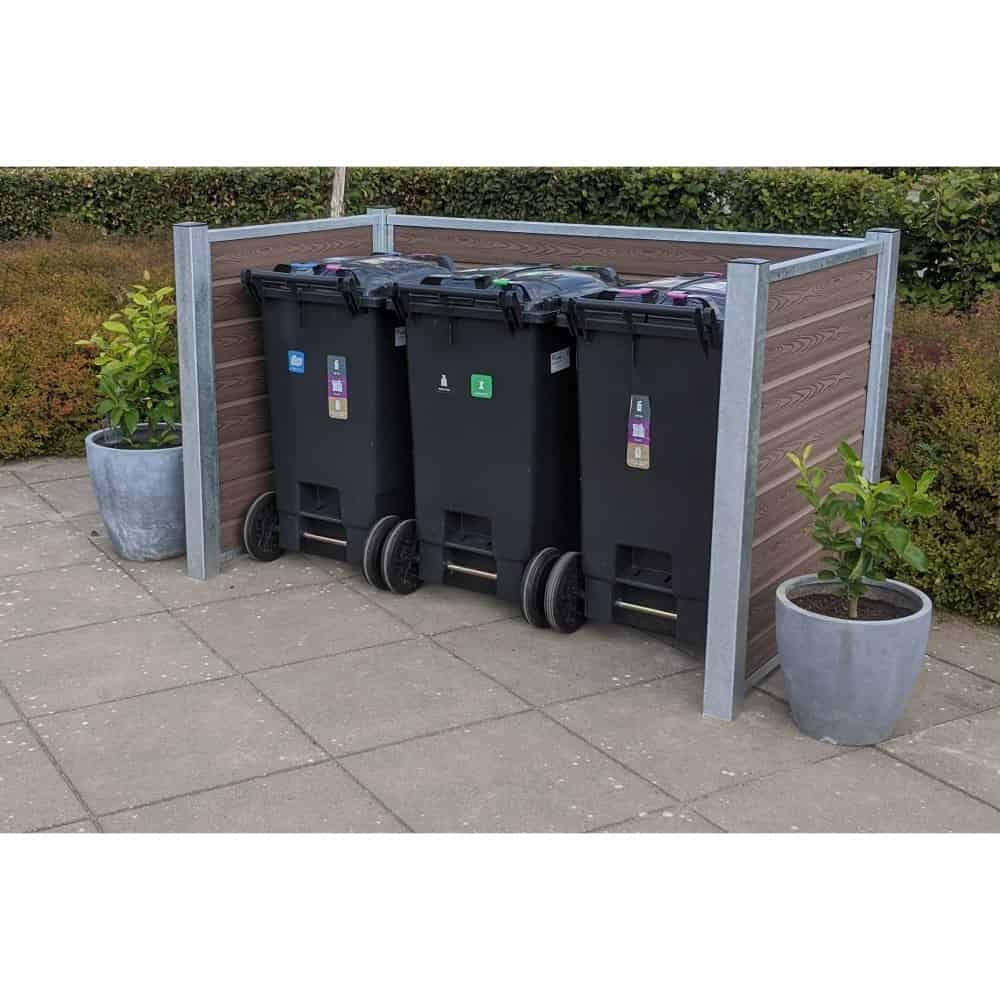 Et affaldshåndteringsområde med tre sorte skraldespande på hjul er omsluttet af et beskedent plankeværk understøttet af stålstolper. To store grå plantepotter med grønt løv sidder på hver side af skraldespandene. Rammerne er en udendørs terrasse med en hæk i baggrunden.