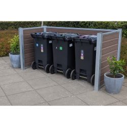 En trio af sorte skraldespande står inden for en Komposit skraldespandsskjul på en asfalteret overflade. Hver beholder har farvede låg og klistermærker, der angiver forskellige typer affald. To potteplanter er placeret på hver side, mens maghoni-buske er synlige i baggrunden.