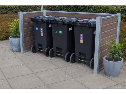 En trio af sorte skraldespande står inden for en Komposit skraldespandsskjul på en asfalteret overflade. Hver beholder har farvede låg og klistermærker, der angiver forskellige typer affald. To potteplanter er placeret på hver side, mens maghoni-buske er synlige i baggrunden.
