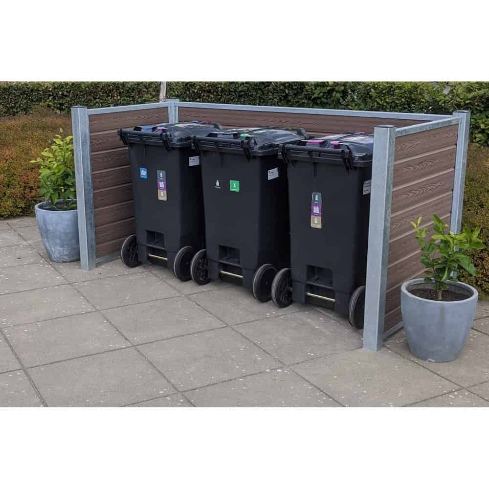 En trio af sorte skraldespande står inden for en Komposit skraldespandsskjul på en asfalteret overflade. Hver beholder har farvede låg og klistermærker, der angiver forskellige typer affald. To potteplanter er placeret på hver side, mens maghoni-buske er synlige i baggrunden.
