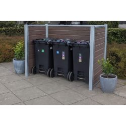 Tre sorte skraldespande på hjul, hver mærket med forskellige genbrugssymboler, er placeret side om side i en komposit skraldespandsskjul med vandrette trælameller og maghoni-detaljer. To potteplanter sidder på fliserne ved siden af skraldespandene med en pænt trimmet hæk i baggrunden.