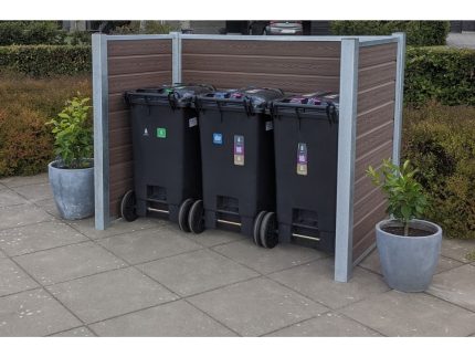 Tre sorte skraldespande på hjul, hver mærket med forskellige genbrugssymboler, er placeret side om side i en komposit skraldespandsskjul med vandrette trælameller og maghoni-detaljer. To potteplanter sidder på fliserne ved siden af skraldespandene med en pænt trimmet hæk i baggrunden.