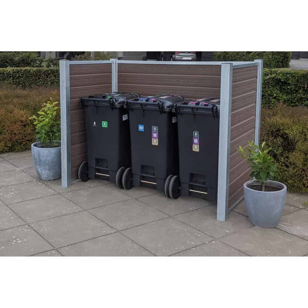 Tre sorte skraldespande på hjul, hver mærket med forskellige genbrugssymboler, er placeret side om side i en komposit skraldespandsskjul med vandrette trælameller og maghoni-detaljer. To potteplanter sidder på fliserne ved siden af skraldespandene med en pænt trimmet hæk i baggrunden.