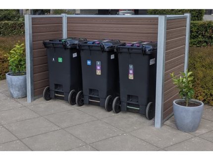 Tre sorte skraldespande på hjul, hver med forskellige farvede etiketter til genbrug, er placeret side om side på et asfalteret område. Beholderne er bakket op af en Komposit skraldespandsskjul med stålstolper og Maghoni paneler. To potteplanter er placeret på hver side af indhegningen.