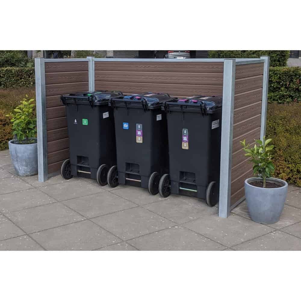 Tre sorte skraldespande på hjul, hver med forskellige farvede etiketter til genbrug, er placeret side om side på et asfalteret område. Beholderne er bakket op af en Komposit skraldespandsskjul med stålstolper og Maghoni paneler. To potteplanter er placeret på hver side af indhegningen.
