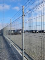 En parkeringsplads med talrige parkerede biler ses gennem et højt Panel Hegn SMART 3D XL 5/5 mm 3000mm - Varmgalvaniseret metalhegn toppet med pigtråd. Himlen er delvist overskyet, og en vindmølle er synlig i det fjerne. Jorden er belagt med beton, og en grussti løber langs hegnet.