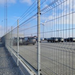 En parkeringsplads med talrige parkerede biler ses gennem et højt Panel Hegn SMART 3D XL 5/5 mm 3000mm - Varmgalvaniseret metalhegn toppet med pigtråd. Himlen er delvist overskyet, og en vindmølle er synlig i det fjerne. Jorden er belagt med beton, og en grussti løber langs hegnet.