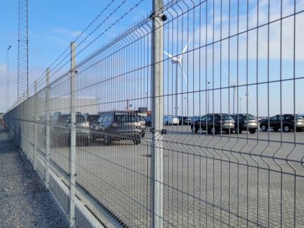 En parkeringsplads med talrige parkerede biler ses gennem et højt Panel Hegn SMART 3D XL 5/5 mm 3000mm - Varmgalvaniseret metalhegn toppet med pigtråd. Himlen er delvist overskyet, og en vindmølle er synlig i det fjerne. Jorden er belagt med beton, og en grussti løber langs hegnet.