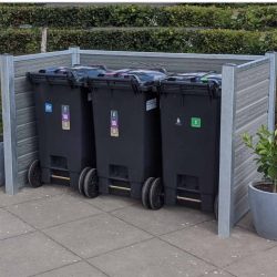 Tre sorte skraldespande på hjul er organiseret i en komposit skraldespandsskjul med robuste stålstolper og en grøn hæk i baggrunden. Hver skraldespand, der er mærket til genbrug, almindeligt affald og komposterbart materiale, er sat mod den slanke Grå-indhegning. To potteplanter flankerer beholderne på hver side.