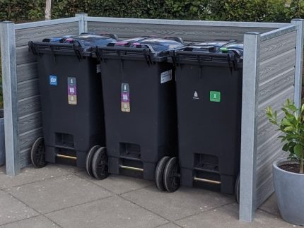 Tre sorte skraldespande på hjul er organiseret i en komposit skraldespandsskjul med robuste stålstolper og en grøn hæk i baggrunden. Hver skraldespand, der er mærket til genbrug, almindeligt affald og komposterbart materiale, er sat mod den slanke Grå-indhegning. To potteplanter flankerer beholderne på hver side.