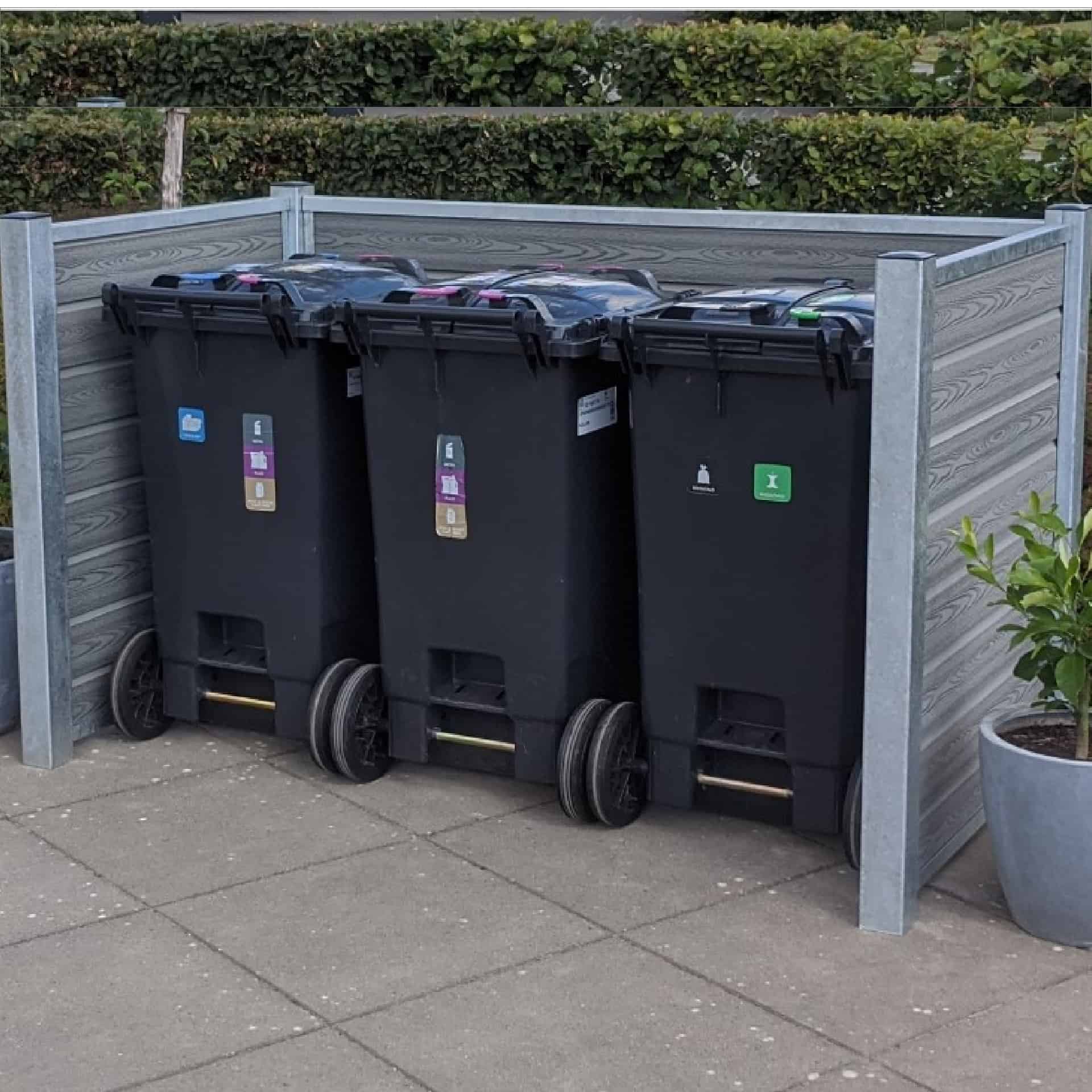 Tre sorte skraldespande på hjul er organiseret i en komposit skraldespandsskjul med robuste stålstolper og en grøn hæk i baggrunden. Hver skraldespand, der er mærket til genbrug, almindeligt affald og komposterbart materiale, er sat mod den slanke Grå-indhegning. To potteplanter flankerer beholderne på hver side.