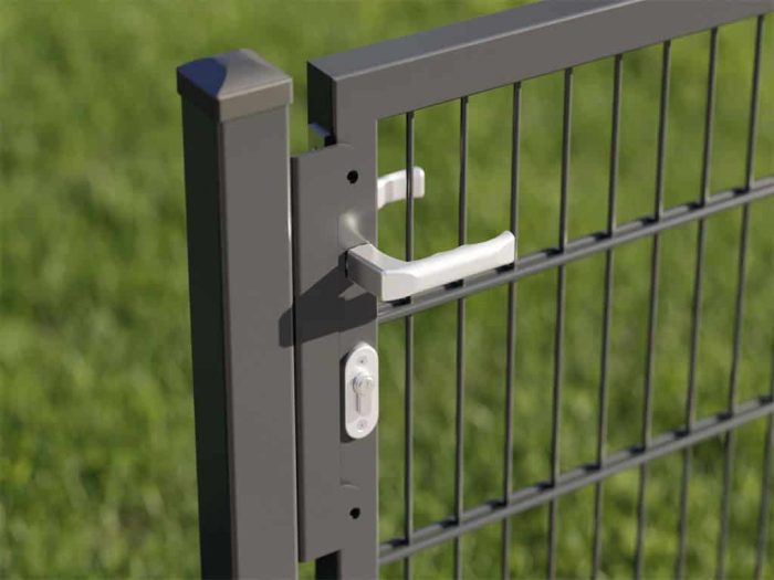 Et nærbillede af en metalportlås fastgjort til en antracitfarvet metalhegnspæl fra Havelåge "BALUSTER"-kollektionen, med både lodrette og vandrette stænger. Det hvide låsehåndtag står i kontrast til den mørke finish, mens den slørede baggrund viser grønt græs.