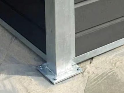 En metalstolpe, der er fastgjort til en betonbase med bolte, står ved siden af mørke vandrette facadebeklædninger og belægningssten - perfekt til at installere Komposit skraldespandsskjul - inkl. stålstolper, stolpehat, top & bund skinner i stål.