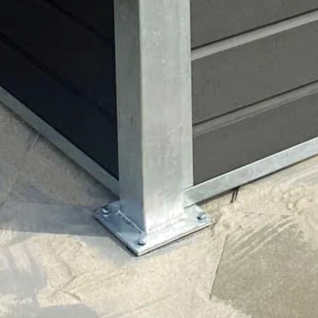 En metalstolpe, der er fastgjort til en betonbase med bolte, står ved siden af mørke vandrette facadebeklædninger og belægningssten - perfekt til at installere Komposit skraldespandsskjul - inkl. stålstolper, stolpehat, top & bund skinner i stål.