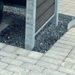 A Komposit skraldespandsskjul inkl. stålstolper, stolpehat, top & bund skinner i stål til 240L står på gråt grus ved siden af en brolagt sti, med benene i gruset og et buet metalstykke på højre side.