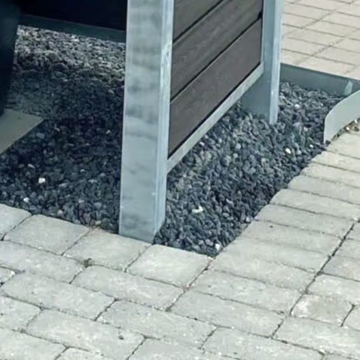 A Komposit skraldespandsskjul inkl. stålstolper, stolpehat, top & bund skinner i stål til 240L står på gråt grus ved siden af en brolagt sti, med benene i gruset og et buet metalstykke på højre side.