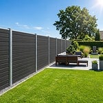 En moderne baggård med et mørkt komposit hegn med stålstolper, grøn græsplæne, siddepladser på terrassen med grå hynder, potteplanter og træer under en klar blå himmel.