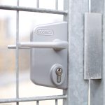 Et nærbillede af en sølv Locinox dørlås sat på en metaltrådport. Låsen har et håndtag på den ene side og et nøglehul på den anden, integreret i 2D Panellåge "TREND" Galvaniseret/Pulverlakeret. Den omgivende portramme og mesh er metallisk og har et industrielt udseende.