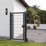 Et moderne sort 2D Panellåge "TREND" står ved siden af et hvidt murstenshus. Porten åbner til et gårdhaveområde prydet med potteplanter og en festlig cirkulær krans med gule bånd hængende på hegnet. Gården kan prale af grønt græs, omgivet af frodige grønne områder.