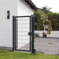 Et moderne sort 2D Panellåge "TREND" står ved siden af et hvidt murstenshus. Porten åbner til et gårdhaveområde prydet med potteplanter og en festlig cirkulær krans med gule bånd hængende på hegnet. Gården kan prale af grønt græs, omgivet af frodige grønne områder.