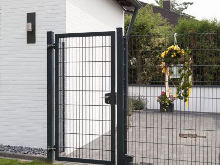 Et moderne sort 2D Panellåge "TREND" står ved siden af et hvidt murstenshus. Porten åbner til et gårdhaveområde prydet med potteplanter og en festlig cirkulær krans med gule bånd hængende på hegnet. Gården kan prale af grønt græs, omgivet af frodige grønne områder.