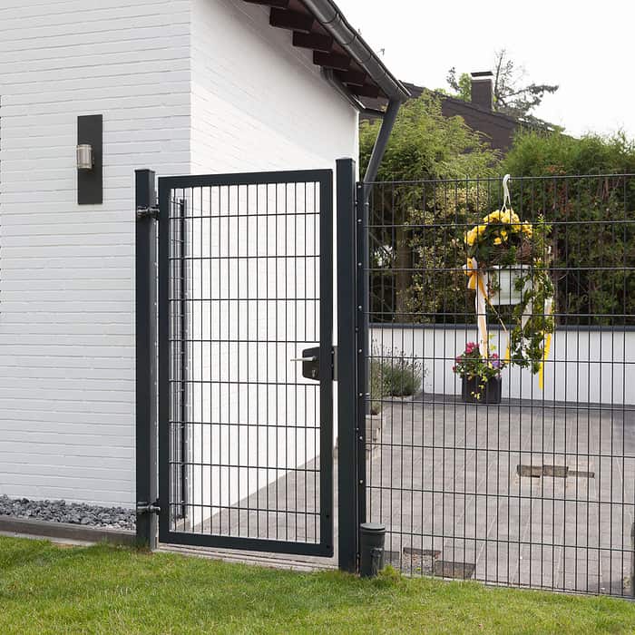 Et moderne sort 2D Panellåge "TREND" står ved siden af et hvidt murstenshus. Porten åbner til et gårdhaveområde prydet med potteplanter og en festlig cirkulær krans med gule bånd hængende på hegnet. Gården kan prale af grønt græs, omgivet af frodige grønne områder.