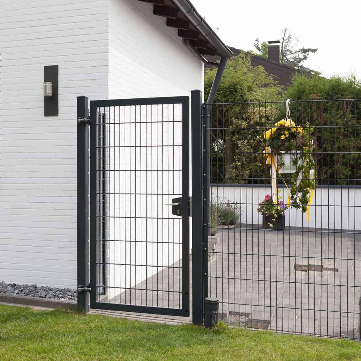 Et moderne sort 2D Panellåge "TREND" står ved siden af et hvidt murstenshus. Porten åbner til et gårdhaveområde prydet med potteplanter og en festlig cirkulær krans med gule bånd hængende på hegnet. Gården kan prale af grønt græs, omgivet af frodige grønne områder.