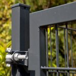 Nærbillede af et sort metalporthængsel med bolte, der fastgør den rektangulære stålramme på 2D Panellåge "TREND" Galvaniseret/Pulverlakeret til en stolpe. Panelet er perfekt justeret, mens baggrunden har sløret løv og naturligt grønt.