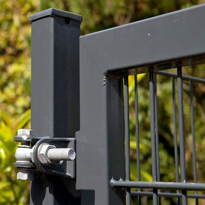 Nærbillede af et sort metalporthængsel med bolte, der fastgør den rektangulære stålramme på 2D Panellåge "TREND" Galvaniseret/Pulverlakeret til en stolpe. Panelet er perfekt justeret, mens baggrunden har sløret løv og naturligt grønt.