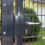 Et nærbillede af 2D Panellåge "TREND" Galvaniseret/Pulverlakeret, med en sort metallåge med sølvhåndtag og låsemekanisme. Baggrunden viser sløret grønt, hvilket tyder på udendørs omgivelser. Denne port ser ud til at være en del af et hegn lavet af lodrette og vandrette metalstænger, hvilket forbedrer dets moderne låge-design.