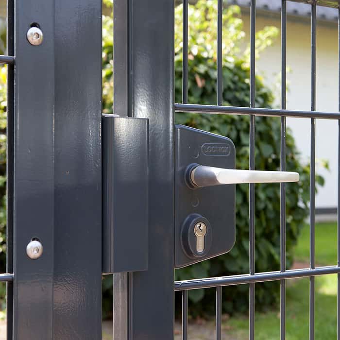 Et nærbillede af 2D Panellåge "TREND" Galvaniseret/Pulverlakeret, med en sort metallåge med sølvhåndtag og låsemekanisme. Baggrunden viser sløret grønt, hvilket tyder på udendørs omgivelser. Denne port ser ud til at være en del af et hegn lavet af lodrette og vandrette metalstænger, hvilket forbedrer dets moderne låge-design.