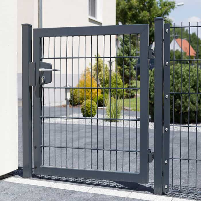 En moderne grå 2D Panellåge "TREND" galvaniseret og pulverlakeret port med lodrette og vandrette sprosser er en del af et hegn i et boligområde. Lågen er lukket og sikret med en lås. Bag porten er der anlagt landskab med grønne buske og små træer. En asfalteret gangbro er foran.