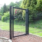 En sort "TREND" 2D Panellåge port, galvaniseret og pulverlakeret, er en del af en indhegnet indhegning i en have. Hegnet og lågen er lavet af trådnetpaneler. Porten står lidt på klem og afslører en grøn, velholdt græsplæne og forskellige buske og træer i baggrunden. Jorden er dækket af små røde småsten.