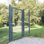 En sort metallåge og hegn med 2D Panellåge "TREND" Galvaniseret/Pulverlakeret står åbent på en grussti, der fører ind i en anlagt have omgivet af træer. De lodrette og vandrette bjælker afslører glimt af græs gennem dem. Området er badet i sollys, hvilket skaber rolige, parklignende omgivelser.