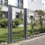 En moderne metalport med lodrette stænger, 2D Panellåge "TREND" Galvaniseret/Pulverlakeret, er en del af et hegn, der omkranser en velholdt have med grønt græs, buske og en bygning med altaner i baggrunden. Haven er delvist skyggefuld af en stor grøn paraply.