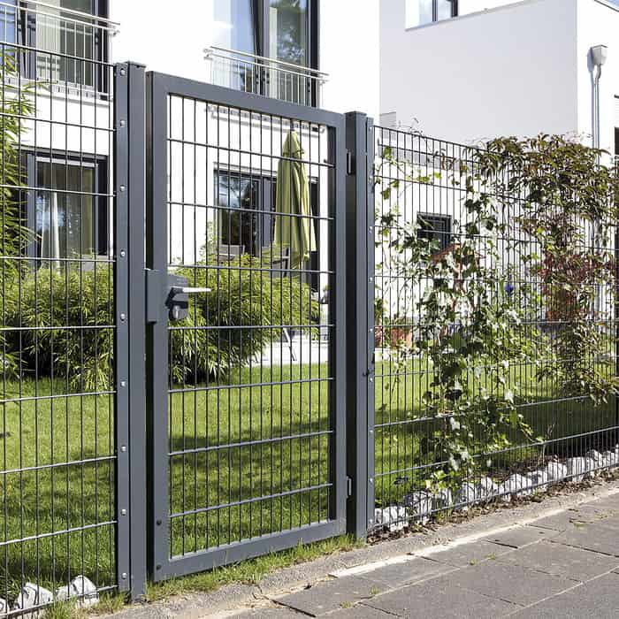 En moderne metalport med lodrette stænger, 2D Panellåge "TREND" Galvaniseret/Pulverlakeret, er en del af et hegn, der omkranser en velholdt have med grønt græs, buske og en bygning med altaner i baggrunden. Haven er delvist skyggefuld af en stor grøn paraply.