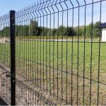 Et Metal Hegn 3D Panel "HF-BUDGET SERIES" Inkl. Stålstolper & Fittings - Pulverlakeret Sorter med lodrette og vandrette bjælker langs omkredsen af en græsmark. I baggrunden er der træer og en hvid bygning med mørkt tag. Himlen er klar og blå, hvilket indikerer en solskinsdag.