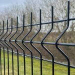 Nærbillede af Metal Hegn 3D Panel "HF-BUDGET SERIES" inkl. stålstolper & beslag i pulverlakeret sort, med et karakteristisk zigzag design. Baggrunden er let sløret og viser bladløse træer og grønt græs, hvilket tyder på en park eller naturlige omgivelser. Den overskyede himmel tilføjer en dæmpet tone til scenen.