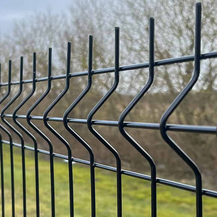 Nærbillede af Metal Hegn 3D Panel "HF-BUDGET SERIES" inkl. stålstolper & beslag i pulverlakeret sort, med et karakteristisk zigzag design. Baggrunden er let sløret og viser bladløse træer og grønt græs, hvilket tyder på en park eller naturlige omgivelser. Den overskyede himmel tilføjer en dæmpet tone til scenen.