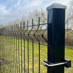 Et nærbillede af Metal Hegn 3D Panel "HF-BUDGET SERIES" Inkl. Stålstolper & Fittings - Pulverlakeret Sort, der viser sit sorte zigzag-mønster, robuste sorte stolpe og støttende klemme. Baggrunden viser græs og bladløse træer under en delvist overskyet himmel.