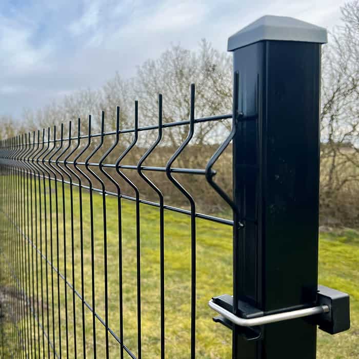 Et nærbillede af Metal Hegn 3D Panel "HF-BUDGET SERIES" Inkl. Stålstolper & Fittings - Pulverlakeret Sort, der viser sit sorte zigzag-mønster, robuste sorte stolpe og støttende klemme. Baggrunden viser græs og bladløse træer under en delvist overskyet himmel.